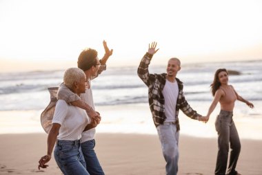 Gün batımında kumlu bir sahilde yürüyen ve gülen dört farklı genç arkadaş, el sallıyor, sıcak ışık ve dalgalarla deniz kenarında kaygısız yaz tatillerinin tadını çıkarıyor.