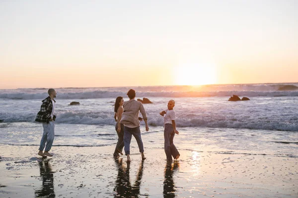 Genç arkadaşlar kumlu bir plajda canlı bir altın saat günbatımının tadını çıkarıyorlar, çıplak ayakla gülüyorlar ve kaygısız bir yaz kaçamağı sırasında ılık okyanus dalgalarında yürüyüp oynuyorlar.