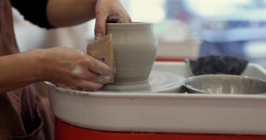 Kadınların elleri, yaratıcı bir atölyede çömlekçi çarkının üzerinde dönen kilden bir tencereyi şekillendirmek için bir alet kullanıyor. Geleneksel zanaatkarlık ve seramik sanatını sergiliyor.