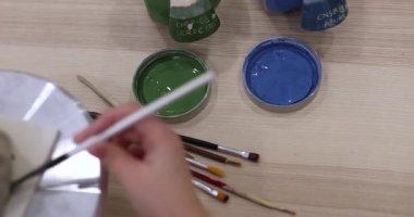 Zanaatkârın eli fırçayı yeşil jöleli bir kaba batırıyor. Çömlekçilik atölyesinde el yapımı seramik bir parça boyamaya hazırlanıyorum. Ahşap masada çeşitli fırçalar ve mavi cila görünür.