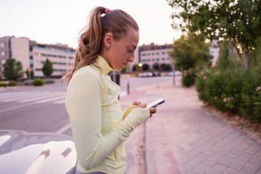 Genç bir kadın şehir caddesinde duruyor, akıllı telefonunu kontrol ediyor ve spor kıyafetlerini giyerken dijital bağlantı kuruyor, modern iletişimi ve şehir yaşam tarzını sembolize ediyor.