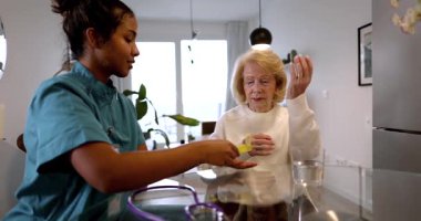 Mutfak masasında oturan yaşlı bir hastaya günlük ilaç veren evde bakım hemşiresi. Profesyonel bakıcı yaşlı bir kadına tedavi için reçeteli ilaçlarını vermesine yardım ediyor.