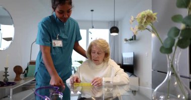 Şefkatli bakıcı, yaşlı bir kadına günlük ilaç tedavisinde yardım ediyor, ilaç takvimini açıklıyor ve rahat bir ev ortamında sağlık yönetimine destek veriyor.