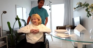 Tekerlekli sandalyedeki gülümseyen yaşlı bir kadına yardım eden, modern, parlak bir dairede yemek yemek için onu yemek masasına iten, yaşlılara ilgi ve destek gösteren,