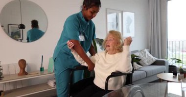 Genç bir kadın sağlık uzmanı oturma odasında kol ve omuz rehabilitasyonu egzersizleri yapan tekerlekli sandalyedeki yaşlı bir kadına şefkatle yardım ediyor. Hareket kabiliyetini ve iyileşmesini teşvik ediyor.