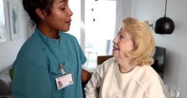 Şefkatli bir sağlık uzmanı huzurevinde gülümseyen yaşlı bir kadına rahatlık ve destek sağlıyor, bakıcı ve hasta arasında sıcak ve şefkatli bir atmosfer sağlıyor.