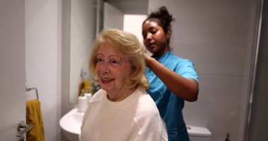 Genç bayan hemşire banyoda yaşlı bir kadının saçını tarayıp şekillendirerek bakım evinde şefkat, yardım ve destek göstererek kişisel bakım sağlıyor.