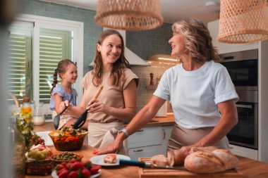 Üç kuşak kadın, büyükanne, anne ve genç kız, neşeli bir şekilde modern bir mutfakta sağlıklı bir yemek pişiriyorlar, mutlu anları paylaşıyorlar ve aile bağlarını paylaşıyorlar.
