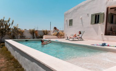 Aile fertleri sakin bir yüzme havuzunda eğlenirken, bir çocuk suya atlarken baba yakalar ve anne güneşli bir günde şezlongda dinlenir.