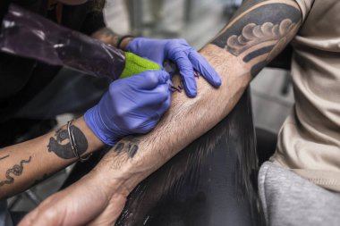 Profesyonel dövme sanatçısı koruyucu eldiven takıyor ve bir stüdyoda müşterinin koluna yeni bir daimi tasarım uygulayan bir makine kullanıyor. Kendini ifade etmeyi ve kişisel tarzını simgeliyor.