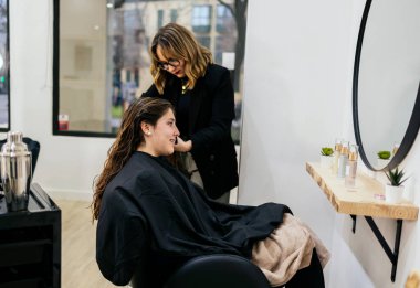 Profesyonel bir kadın tasarımcı, genç bir kadının uzun saçlarını parlak ve modern bir salonda kesiyor. Doğal ışık ile arkadaşça bir randevu sırasında yeni ve şık bir görünüm yaratıyor.