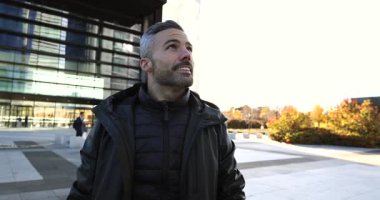 Modern şehir meydanında dikilen sakallı, gri saçlı şık şehirli adam yüzünde hafif bir gülümsemeyle, güneşli günün tadını çıkartıyor.