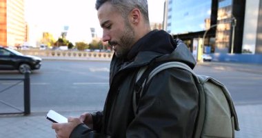 Orta yaşlı, gri saçlı ve sırt çantalı bir adam işlek bir caddede yürürken akıllı telefonuna bakıyor, güneşli bir günde sosyal medyada mesajlaşırken ya da gezinirken gülümsüyor.