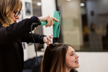 Modern bir kuaförde genç bir kadının uzun saçlarını fön makinesiyle kurutup şekillendiren profesyonel bir kuaför. Yeni bir görünüş ve ilgili müşteri hizmetleri ortamı yaratıyor.