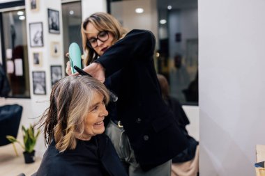 Profesyonel kadın kuaför gülümseyen olgun bir müşteriye saç bakım hizmeti veriyor, saç kurutma makinesi kullanıyor ve saçını bir güzellik salonunda tarıyor.