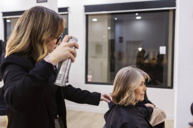 Kuaför, olgun bir kadının kısa saç modeline saç spreyi sıkıyor. Modern bir kuaför salonunda profesyonel bakım ve yeni bir tarz sağlıyor.