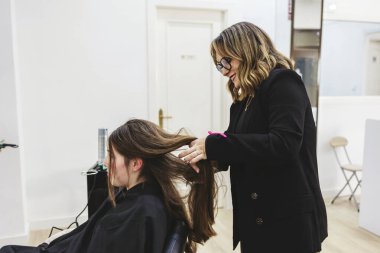 Profesyonel kuaför genç bir kadının uzun kahverengi saçlarını dikkatlice şekillendiriyor, modern bir kuaförde güzellik hizmeti veriyor, saç bakımına ve yeni bir görünüme odaklanıyor.