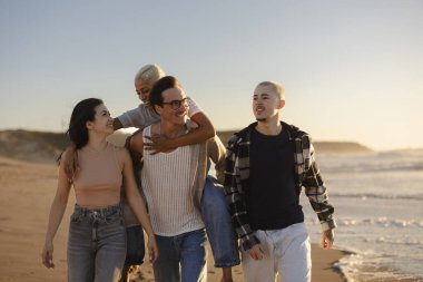 Altın saat boyunca kumlu bir sahilde neşeyle yürüyen bir grup yetişkin arkadaş, bir kişi sırtına biniyor, hepsi gülümsüyor ve bir anlık mutluluğu paylaşıyor.