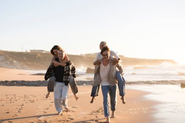 İki neşeli genç çift, kumsalda çıplak ayakla yürürken partnerlerini sırtlarında taşırlar. Okyanus dalgaları ve güzel bir günbatımının keyfini çıkarırlar. Sıcak, altın bir atmosfer yaratırlar.