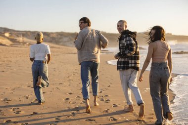 Çeşitli genç arkadaşlar gün batımında kumlu bir plajda birlikte çıplak ayakla koşuyorlar, keyifli bir anı paylaşıyorlar ve yaz tatillerinde dışarıda eğleniyorlar.