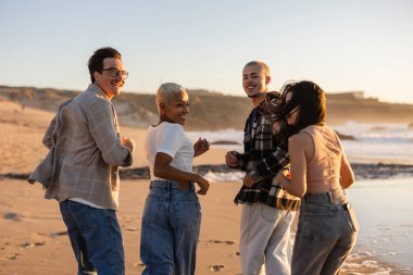 Gün batımında kumlu bir plajda koşarken gülümseyen bir grup genç arkadaş neşeli anlar paylaşıyor ve dışarıda arkadaşlığı kutluyorlar.