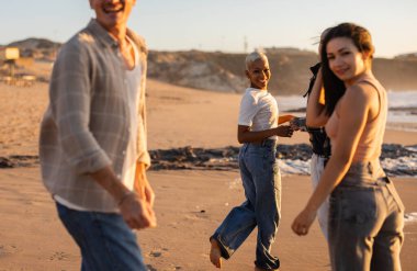 Altın saatinde kumlu bir sahilde çıplak ayakla yürüyen, gülümseyen ve geriye bakan, kaygısız yaz anlarını paylaşan bir grup arkadaş.