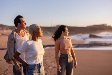 Çeşitli arkadaşlar kumlu bir sahilde huzurlu bir altın saatin tadını çıkarıyorlar, birlikte duruyorlar ve okyanus ufkuna doğru bakıyorlar, arkadaşlığı kucaklıyorlar ve ortak bir huzur anlarını paylaşıyorlar.
