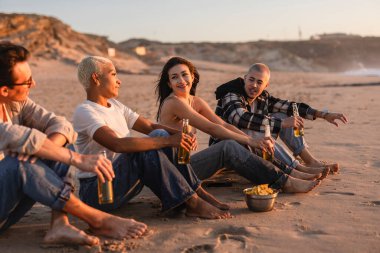 Bir grup farklı yetişkin arkadaş kumsalda altın saat boyunca dinleniyor, kumda oturuyor, şişeleri tutuyor ve abur cubur yiyor, birlikte mutlu bir anı paylaşıyorlar.
