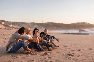 Bir grup farklı genç kumsalda dinleniyor, birlikte içki ve atıştırmalıkların tadını çıkarıyor, öğleden sonra saatlerinde açık hava buluşması yapıyorlar.