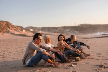 Gün batımında kumlu bir plajda oturup, birlikte içki ve atıştırmalıkların keyfini çıkaran, sosyal bağları, mutluluğu ve yaz rahatlığını gösteren çeşitli arkadaşlar grubu.
