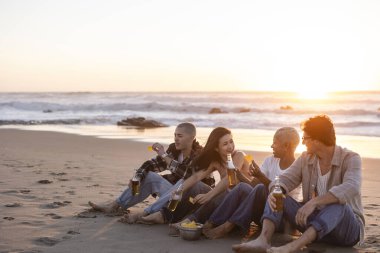 Çeşitli yetişkin arkadaşlar kumlu bir plajda dinleniyor, okyanus kenarında sıcak bir gün batımında sosyalleşirken içecek ve atıştırmalıkların tadını çıkarıyorlar.