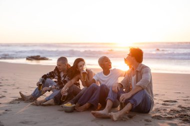 Gün batımında kumlu bir sahilde oturup gülen ve okyanus kenarında sosyalleşirken yiyecek ve içeceklerin tadını çıkaran bir grup yetişkin arkadaş.
