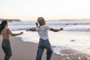 Kumlu bir plajda huzurlu bir gün batımının keyfini çıkaran iki farklı genç kadın okyanus dalgalarına taş atıp seyahat maceraları sırasında neşeli bir aktivite ve özgürlük anlarını paylaşıyorlar.