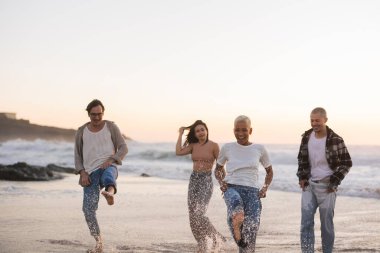 Çeşitli yetişkin arkadaş grupları gün batımında sahilde keyifli bir yürüyüş yapıyorlar, su sıçratıyorlar ve gülüyorlar, yaz özgürlüğünü ve bağlantıyı paylaşıyorlar.