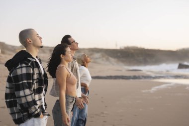 Gün batımında kumlu bir plajda duran, gözleri kapalı derin derin nefes alan, okyanus boyunca doğayla bir anlık bir bağ ve farkındalık yaşayan genç insanlar.