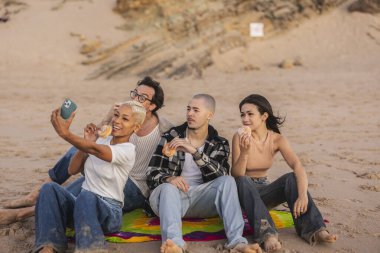 Bir grup genç arkadaş kumlu bir plajda oturup donutlarını paylaşırken ve gülümserken bir kadın akıllı telefon kullanarak rahatça dışarıda geçirdiği anın tadını çıkarıyor.