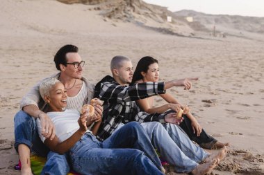 Çeşitli yetişkin arkadaş grupları kumlu bir sahilde dinleniyor, tatlı atıştırmalıkların tadını çıkarıyorlar ve uzaktaki bir şeyi işaret ediyorlar, boş zamanlarını paylaşıyorlar.