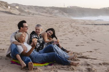 Çeşitli yetişkin arkadaş grupları tatlı hamur işlerinin ve sıcak sohbetin keyfini çıkarırken kumlu bir sahilde keyifli bir toplantının ortasında dinleniyorlar.