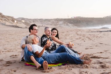 Çeşitli arkadaş grupları güneşli bir plajda gevşeyip gülüyor ve sohbet ederken atıştırmalık ve donut paylaşıyor, boş zamanların, bağ kurmanın ve tatil havasının tadını çıkarıyor.