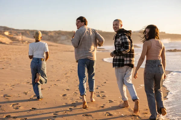 Çeşitli genç arkadaşlar gün batımında kumlu bir plajda birlikte çıplak ayakla koşuyorlar, keyifli bir anı paylaşıyorlar ve yaz tatillerinde dışarıda eğleniyorlar.