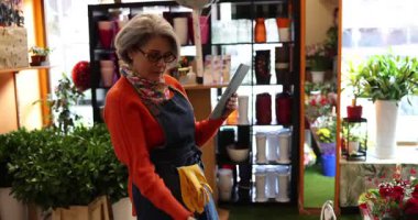 Çiçekçi olarak çalışan olgun bir kadın, dijital tablet kullanarak küçük işletmesinin envanterini ve online siparişlerini düzenliyor, güzel çiçekçisinin içinde kırmızı bir Gerbera papatyası tutuyor.