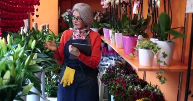 Çiçekçilikte deneyimli bir yönetici. Çiçek ve bitkilerin envanterini dijital tabletle çıkarırken renkli orkideler ve diğer çiçek aranjmanlarıyla dolu rafların arasında duruyor.