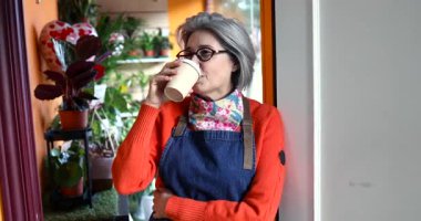 Gri saçlı olgun çiçekçi çiçek dükkanının girişinde beklerken dinlendirici bir kahve molası veriyor. Düşünceli bir şekilde dışarı bakıyor ve işine devam etmeden önce saatini kontrol ediyor.