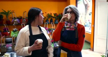 Farklı yaşlarda, önlük giyen neşeli kadın çiçekçiler birlikte kahve molası verip çiçekçinin içinde sohbet ediyorlar. İş arkadaşları çalışma saatlerinde dostça bir anı paylaşıyorlar