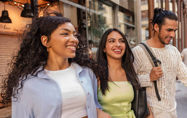 Diverse group of three happy young adults, two women and one man, smiling and enjoying a casual walk along an urban street with shops and buildings in the background