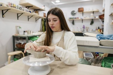 Önlüklü genç bir kadın pikapta küçük bir kase şekillendirirken iyi donanımlı bir zanaatkar atölyesinde seramik kursunda zanaatkarlık geliştiriyor.