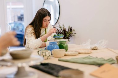 Bir çömlek atölyesinde el yapımı bir seramik kaseyi güzel bir fırçayla boyarken konsantre olan genç bir kadın, bir sanat atölyesi sırasında beceri ve yaratıcılık geliştiriyor.