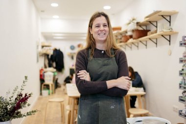 Önlük takan ve kollarını kavuşturan gülümseyen kadın, seramik ve çömlek atölyesinde güvenle duruyor, girişimcilik ve zanaatkarlığı temsil ediyor.