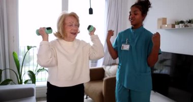 Destekleyici bayan hemşire oturma odasında halter egzersizleriyle neşeli bir son sınıf kadınına yardımcı oluyor. Fizik tedavi ve ev sağlığı hizmetleri yaşlılar için aktif yaşlanma ve rehabilitasyonu teşvik ediyor
