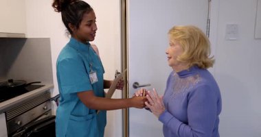 Önlüklü genç bir bayan bakıcı ev ziyareti sırasında yaşlı bir kadının elini ve panosunu tutuyor, güven verici bir şekilde konuşurken aynı zamanda profesyonel tıbbi destek ve konfor sunuyor.